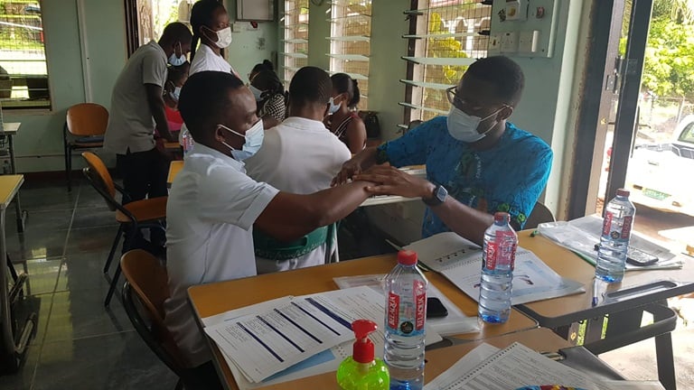 2 learners are sitting at desks in a classroom setting across from each other carrying out a range of motion teat of the wrist. They are wearing masks, and have instructions on the desk in front of them